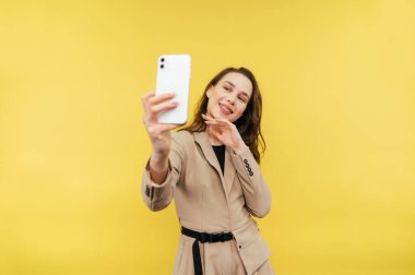 Ceketli mutlu bir kadın sarı arka planda özçekim yapıyor yüzünde gülümsemeyle akıllı telefondaki kameraya bakıyor. İzole edilmiş