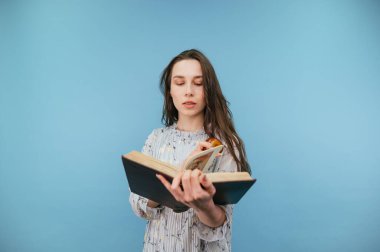 Elbiseli çekici esmer ciddi bir yüzle mavi arka planda bir kitap okuyor ve elinde bir elma tutuyor. İzole edilmiş.