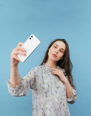 Elbise giymiş çekici romantik bir kadın mavi arka plandaki akıllı bir telefondan fotoğraf çekip kameraya poz veriyor. dikey.