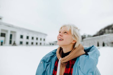 Sarı saçlı ve kalın elbiseli mutlu bir sarışın karla kaplı bir arka planda duruyor ve yüzünde bir gülümsemeyle yukarı bakıyor..