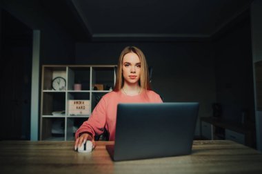 Konsantre olmuş genç bir kız evinde dizüstü bilgisayar başında oturuyor ve bilgisayardan ders çalışıyor, ciddi bir yüzle kameraya bakıyor..