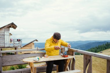 Erkek turist dağlardaki bir bungalovun terasında oturur ve arka plandaki güzel manzaralı ocakta yemek yemeye hazırlanır..
