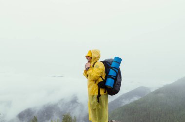 Sırtında sisli manzaralı bir sırt çantasıyla dağın tepesinde duran sarı yağmurluklu bir erkek turistin fotoğrafı..