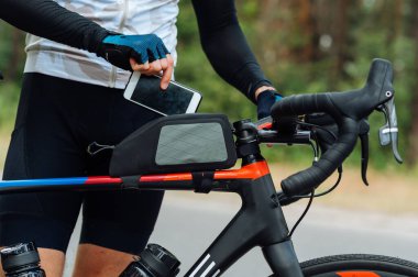 Sportif kıyafetli bir erkek bisikletçi bisikletinin çerçevesinde bir miktar olan akıllı bir telefon koyar. Boş vakitte bisiklet, hobi.