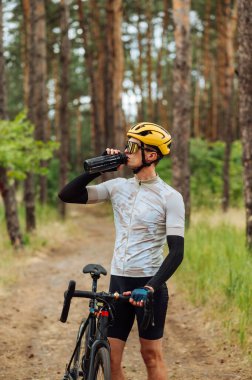 Erkek bisikletçi ormanda güzel doğal manzaralı bir arka planda bisikletle durur ve bir şişeden su içer..