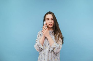 Elbiseli romantik kadın telefonda mavi arka planda konuşuyor, yüzünü çeviriyor ve gülümsüyor..
