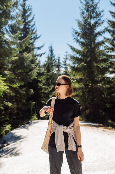 Attractive female tourist in casual clothes stands in the mountain forest in the snow in light summer clothes and in sunglasses and looks away. Walking in the mountains