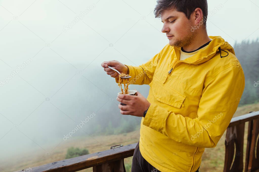 Male tourist eating vermicelli from a cup in the mountains on a background of misty forest, standing on the terrace of a wooden house.