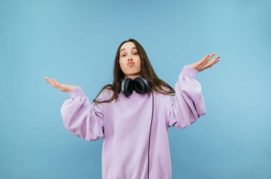 Mor kazaklı şaşkın kadın mavi arka planda duruyor ve şaşkın bir yüzle kameraya bakıyor ve kollarını açıyor..