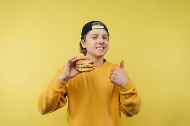 Pozitif genç bir adam elinde bir hamburgerle sarı bir arka planda duruyor, yüzünde bir gülümsemeyle kameraya bakıyor ve sevdiği bir jesti gösteriyor..