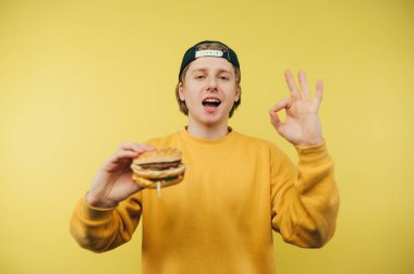 Şapkalı ve kazaklı neşeli genç adam elinde bir hamburgerle sarı bir arka planda duruyor, yüzünde bir gülümsemeyle kameraya bakıyor, elinde bir hamburger tutuyor ve bir jest yapıyor.