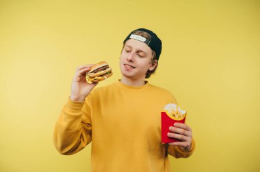 Pozitif genç adam sarı arka planda hamburger ve patates kızartması ile duruyor ve aç bir bakışla fast food 'a bakıyor ve yüzünde bir gülümseme var.