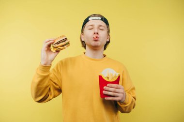 Sıradan kıyafetler içindeki komik adam sarı arka planda fast food yiyor, elinde bir burger tutuyor ve patates kızartması yiyor..