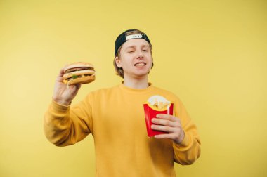 Şapkalı ve sarı kazaklı mutlu genç adam sarı bir arka planda elinde patates kızartması ve bir burgerle duruyor ve yüzünde bir gülümsemeyle kameraya bakıyor..