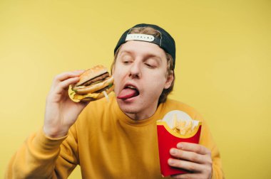 Şapkalı ve sarı kazaklı mutlu genç adam sarı bir arka planda elinde patates kızartması ve bir burgerle duruyor ve yüzünde bir gülümsemeyle kameraya bakıyor..