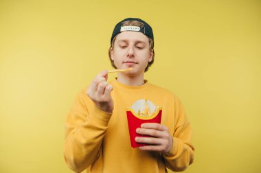 Şapkalı, renkli tişörtlü, sarı arka planda izole edilmiş ellerinde patates kızartması olan yakışıklı bir adam. Genç adam öğle yemeğinde patates kızartması yiyor..
