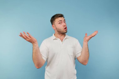 Kafası karışmış genç bir adam, beyaz bir tişört giyiyor, mavi arka planda duruyor ve kollarını karmaşadan uzak tutuyor..