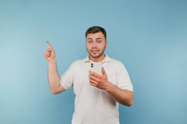 Elinde akıllı telefonuyla tatminsiz genç bir adam mavi bir arka planda duruyor, boş bir alanda parmağını uzaya doğrultuyor ve kameraya dik bir yüzle bakıyor..