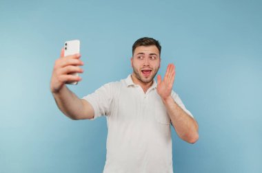 Pozitif adam akıllı telefondan görüntülü iletişim kuruyor. Yüzünde gülümseme var. Mavi arka plandaki kameraya hoş geldin..