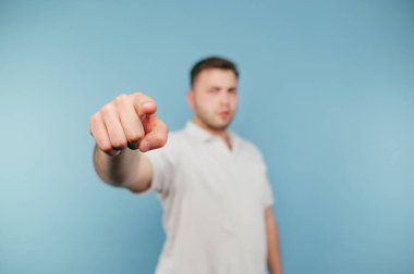 Beyaz tişörtlü bir adam, mavi arka planda kameraya parmağını gösteriyor, parmağının yakın fotoğrafını..