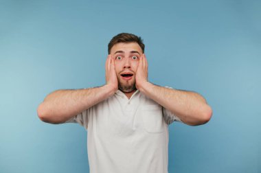 Beyaz tişörtlü, mavi arka planda kılları olan şok olmuş bir adamın portresi, şok içinde ellerini başının arkasına koydu ve kameraya baktı.