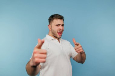 Beyaz tişörtlü, mutlu yüzlü, duygusal pozitifliğe sahip bir adam mavi arka planda izole edilmiş parmaklarını kameraya gösteriyor.