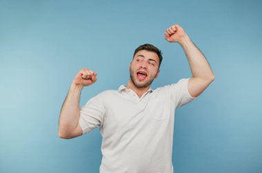 Tüylü ve beyaz tişörtlü neşeli adam mavi arka planda mutlulukla dans ediyor ve gülümsüyor..