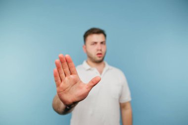 Beyaz tişörtlü ciddi bir adam el hareketini gösteriyor. Dur ve kameraya bakıyor. Tatminsiz bir yüzle, mavi bir arka planda izole edilmiş..