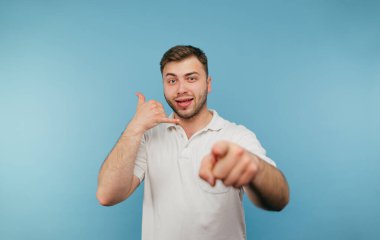 Tüylü olumlu bir adam kamerayı işaret ediyor ve mavi arka planda gülen yüzlü bir jest yapıyor..