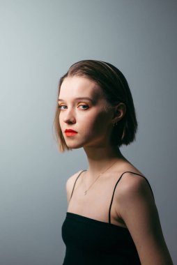 Makyajlı, üstten giyinmiş, gri duvarlı arka planda kameraya poz veren ve ciddi bir yüzle kameraya bakan çekici bir kadın.