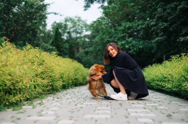 Gülümseyen stil sahibi bir kadın, köpek cinsi Pomeranian Spitz 'le birlikte yeşil bir parkın arka planında yürüyüş yapıyor ve mutlu bir yüzle kameraya bakıyor..