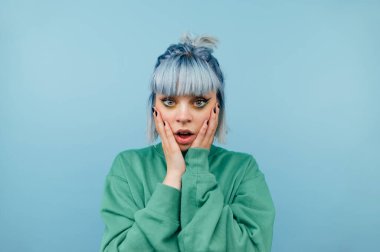 Mavi arka planda renkli saçlı, kameraya bakan, yanaklı, yeşil kazaklı genç bir kızın portresi.