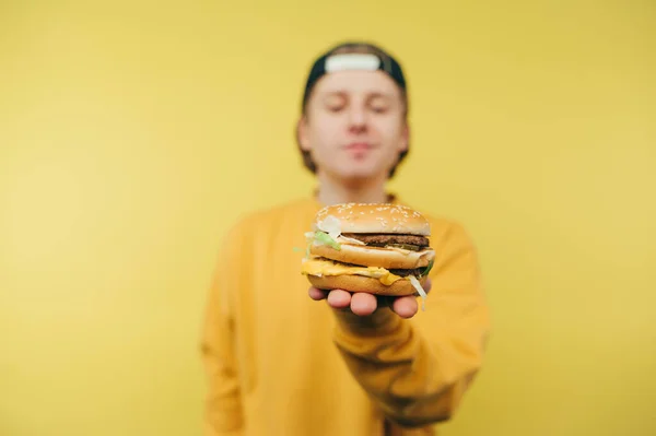 穿着休闲装的积极向上的家伙在镜头前展示黄色背景的大汉堡 脸上挂着笑容 一个年轻人手中拿着一个汉堡包的近照 — 图库照片