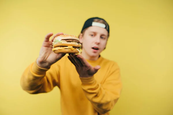 평상복을 모자를 젊은이가 노란색 배경의 햄버거를 맛있게 먹는다 패스트푸드의 — 스톡 사진