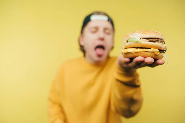 有趣的饥饿的家伙伸出舌头向摄像机展示了一个黄色背景的美味汉堡 — 图库照片