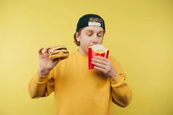 배고픈 남자가 노란색 배경에 감자튀김 햄버거를 패스트푸드를 먹으면서 — 스톡 사진