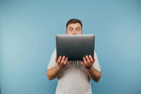 用蓝色背景手拿着笔记本电脑的震惊男人的画像 带着滑稽的感情的眼神看着屏幕 — 图库照片