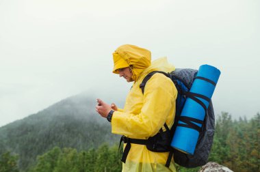 Dağlarda sırtında sırt çantası olan ve yağmurluk giyen bir adam, dağların sisli arka planlarına ve kozalaklı ormanlara karşı ciddi bir yüze sahip bir akıllı telefon kullanıyor..