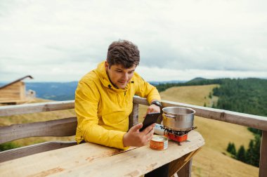 Sarı ceketli bir erkek turist, balkondaki dağlarda oturur, ocakta yemek hazırlar ve arka plandaki güzel manzaralara karşı akıllı telefon kullanır..