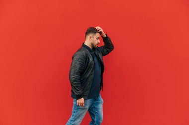 Sakallı ve deri ceketli yetişkin bir adam kırmızı bir duvarın arkasında yürür..