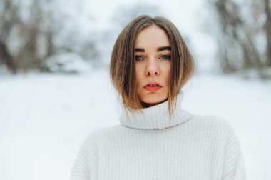 Beyaz kazaklı çekici bir kadın kışın, arka planda, bulanık kar manzaralı bir yerde dikilir ve kameraya bakar..