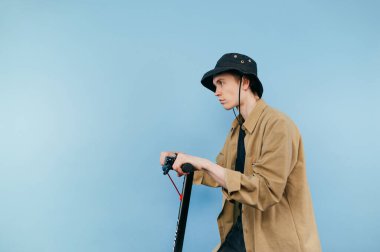 Panama 'lı ciddi bir genç adam mavi arka planda elektrikli bir el scooterına biniyor ve yüzünü çeviriyor. Boşluğu kopyala