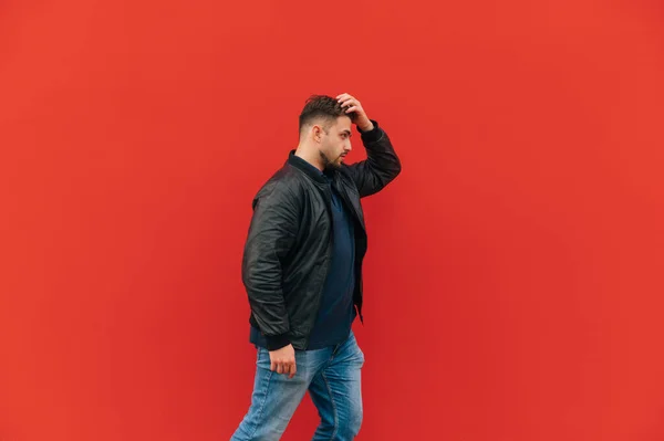 stock image An adult man with a beard and a leather jacket walks against the backdrop of a red wall, side view.