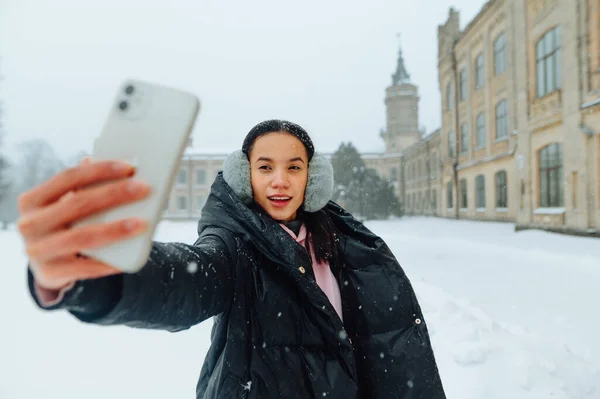 在一个大雪的日子里 女士会在冬天自拍 穿着暖和外套头罩的漂亮女人站在大街上 站在雪地里 拿着智能手机在镜头前自拍 — 图库照片