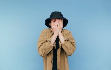 Panamalı korkmuş adam mavi bir arka planda izole edilmiş, elleriyle yüzünü örtüyor ve avuçlarından kameraya bakıyor..