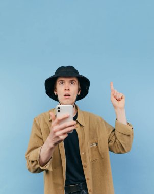 Şok olmuş genç adam, sıradan kıyafetler ve panama giymiş elinde akıllı telefonuyla duruyor ve şaşırmış yüzüyle fotokopi alanını işaret ediyor.