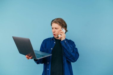 Gömlekli ciddi bir iş adamı dizüstü bilgisayarı mavi arka planda ve telefonda konuşuyor..
