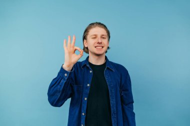 Şık giyinmiş, mavi arka planda tişört giyen neşeli bir adam kameraya bir jest yapıp göz kırpıyor.