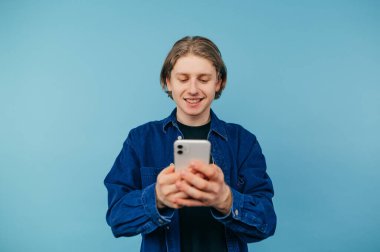 Gömlekli olumlu bir adam elinde akıllı bir telefonla mavi bir arkaplanda duruyor ve yüzünde gülümsemeyle bir mesaj yazıyor..