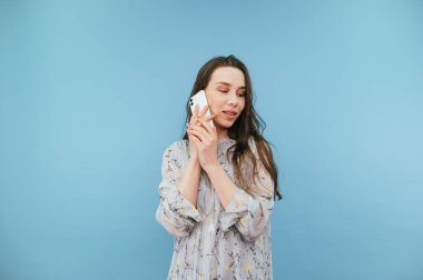 Şirin elbiseli, gülümseyen güzel bir kadın telefonda mavi bir arkaplanda, yüzünde bir gülümsemeyle iletişim kuruyor..